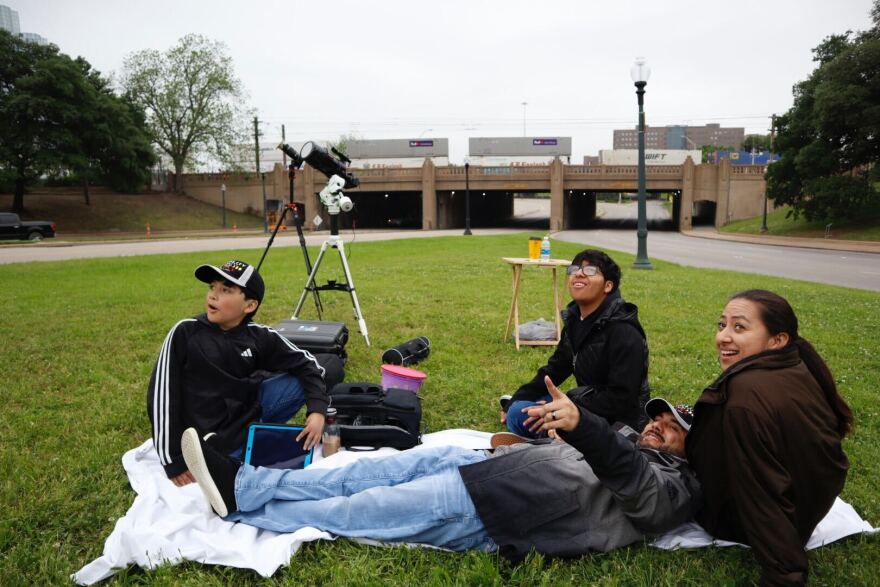 Four people sit on a blanket in the grass looking up toward the left of the frame at the sky. Behind them are two cameras on tripods pointed at the sky.