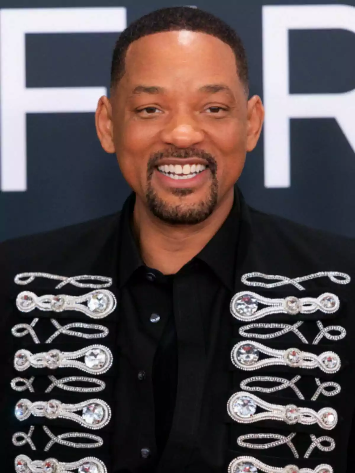Will Smith, smiling with a beard and short hair, wears a black jacket adorned with shiny details. Will Smith, smiling with a beard and short hair, wears a black jacket adorned with shiny details.