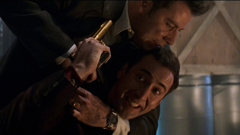 Nicolas Cage as Sean Archer wearing Castor Troy's face and hJohn Travolta as Castor Troy wearing Sean Archer's face, both pointing guns to each other's head in close proximity in Face/Off