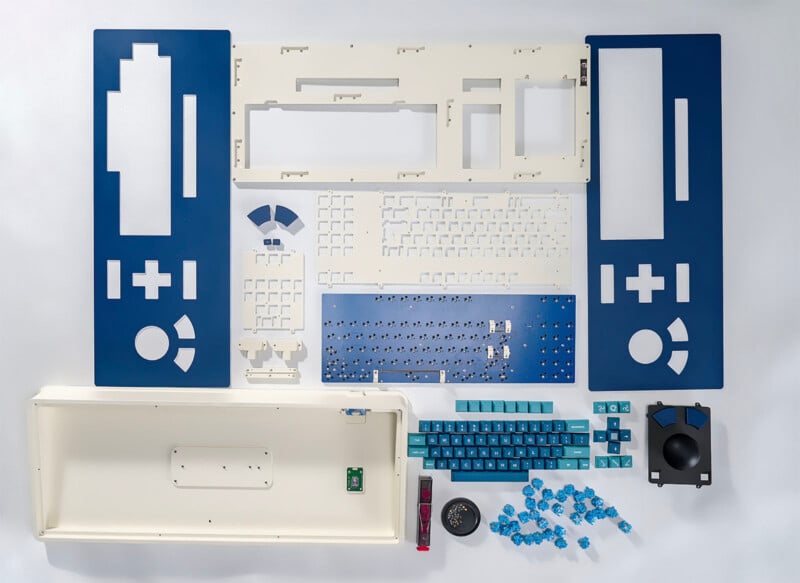 Neatly arranged keyboard parts and components, including white and blue panels, a circuit board, keys, switches, a dial, screws, and other assembly parts, displayed on a white background.