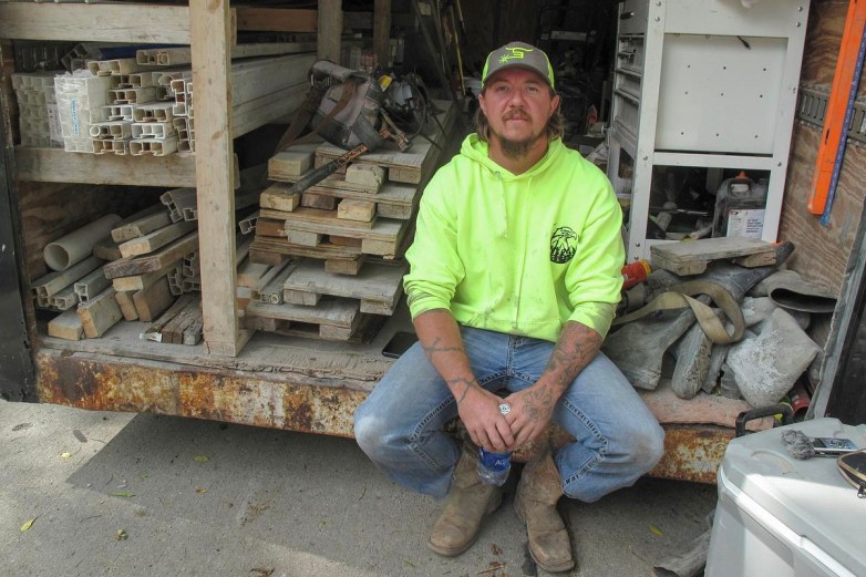 Man in yellow jacket and jeans sits next to lumber and other construction supplies.