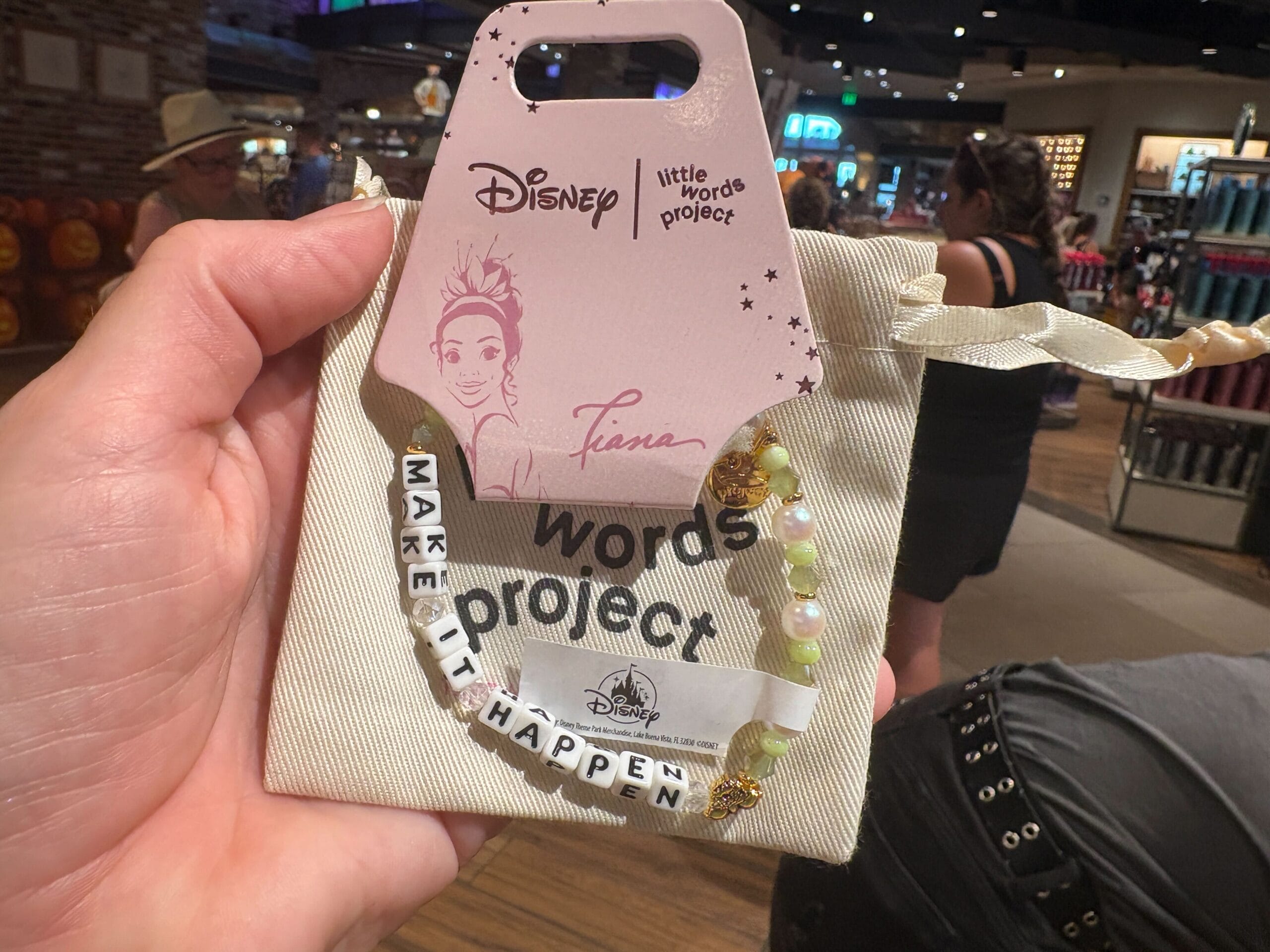 A hand holds a Disney Tiana “Make It Happen” beaded bracelet in a store with people and shelves in the background.