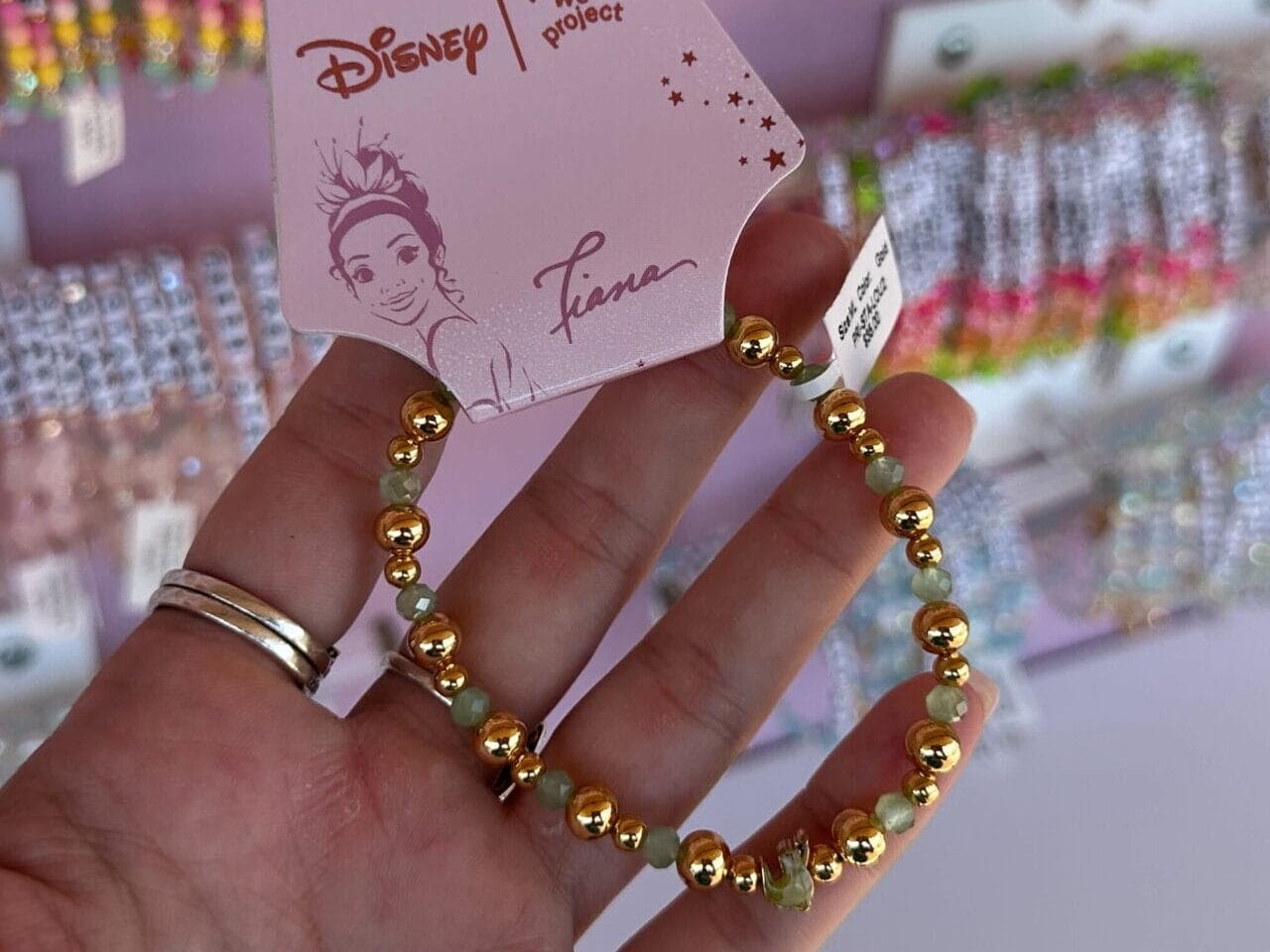 A hand holds a gold beaded Disney Princess Tiana bracelet in front of a display of colorful bracelets.