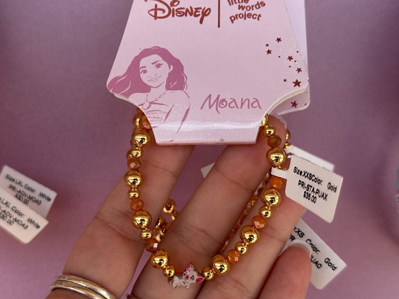 Hand holding a Moana-themed Disney bracelet with gold beads, displayed on a pink background among other beaded bracelets.