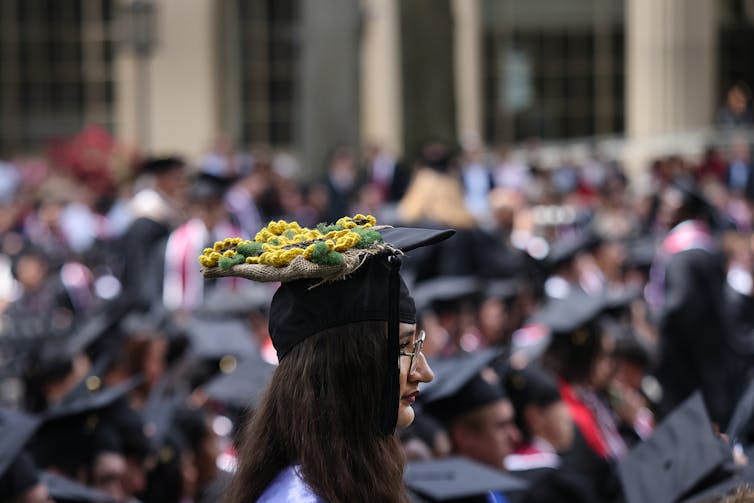 A woman with dark hair and a black graduation cap with yellow flowers is seen in front of a crowd of people seated also wearing black caps.
